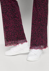 Mustat housut, joissa on rohkea vaaleanpunainen abstrakti kuosi, tähdelliset helmat, yhdistetty valkoisiin lenkkareihin, joissa on sileä pinta ja rento muotoilu.