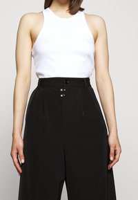 Top blanco sin mangas combinado con pantalones negros de cintura alta que cuentan con cierre de botón y dos pliegues frontales. Tela suave con un diseño ajustado.