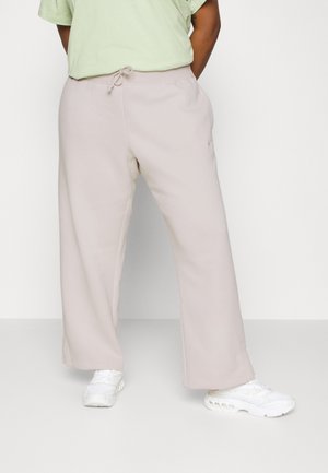 Osoba ubrana w jasnobeżowe spodnie o szerokich nogawkach ze ściągaczem w talii oraz białe trampki, stojąca na tle jednolitego białego tła.