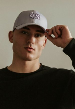 Purple cap with a shell emblem, worn by a person in a black shirt. The cap features a structured design and smooth fabric texture.