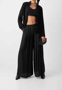 Top corto nero, camicia lunga con bottoni, e pantaloni a gamba larga. Tessuto liscio, vestibilità rilassata. Accessorio: piccola pochette nera. Accessori: gioielli semplici.
