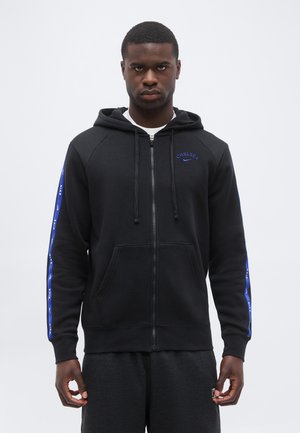 Homme portant un sweat à capuche noir zippé avec les logos bleus "Chelsea" et Nike, debout les mains détendues devant un fond blanc uni.