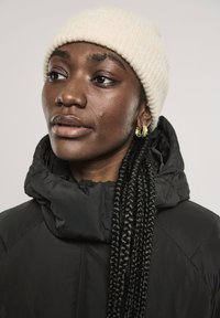 Chaqueta de plumas negra con cuello alto, gorro de punto acanalado crema y pendientes de aro dorados. Se puede ver un peinado trenzado texturizado.