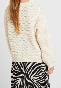 Cremfarbener, strukturierter Strickpullover mit lockerer Passform, gerippten Bündchen und Saum, getragen über einem schwarz-weißen gestreiften, plissierten Rock.