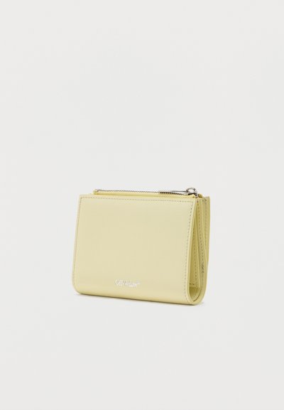 Πορτοφόλι από δέρμα σε εξασθενισμένο κίτρινο χρώμα με κλείσιμο με φερμουάρ και ανάγλυφο λογότυπο Off-White στο μπροστινό μέρος, μινιμαλιστικός σχεδιασμός σε λευκό φόντο.