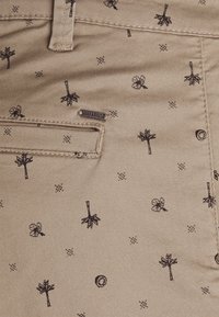 Béžová tkanina s černými vyšívanými vzory stromů, květin a geometrických tvarů. Obsahuje přední kapsu a malou značkovou etiketu.