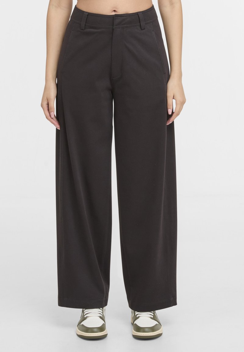 Vue de face d'une personne portant un pantalon noir taille haute à jambes larges et des baskets blanches et vertes, debout sur un fond clair uni.