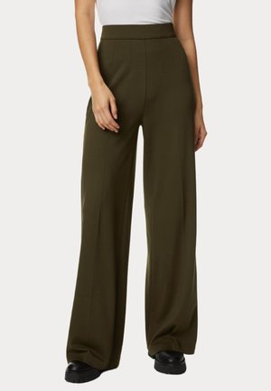 Personne portant un pantalon taille haute olive à jambes larges, un haut blanc sans manches et des chaussures noires, debout devant un fond uni et clair.