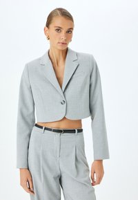 Blazer gris recortado hecho de tela texturizada con solapas muescas, cierre de un solo botón y corte ajustado. Combinado con pantalones de talle alto.