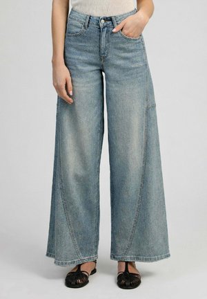 Personne portant un jean bleu délavé taille haute à jambes larges, une main dans la poche, et des sandales plates tissées noires sur un fond clair uni.