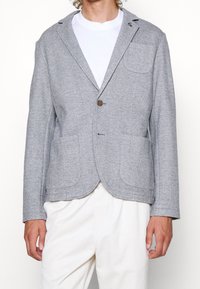 Hombre con blazer gris claro con dos bolsillos frontales y un bolsillo en el pecho sobre una camiseta blanca y pantalones blancos.