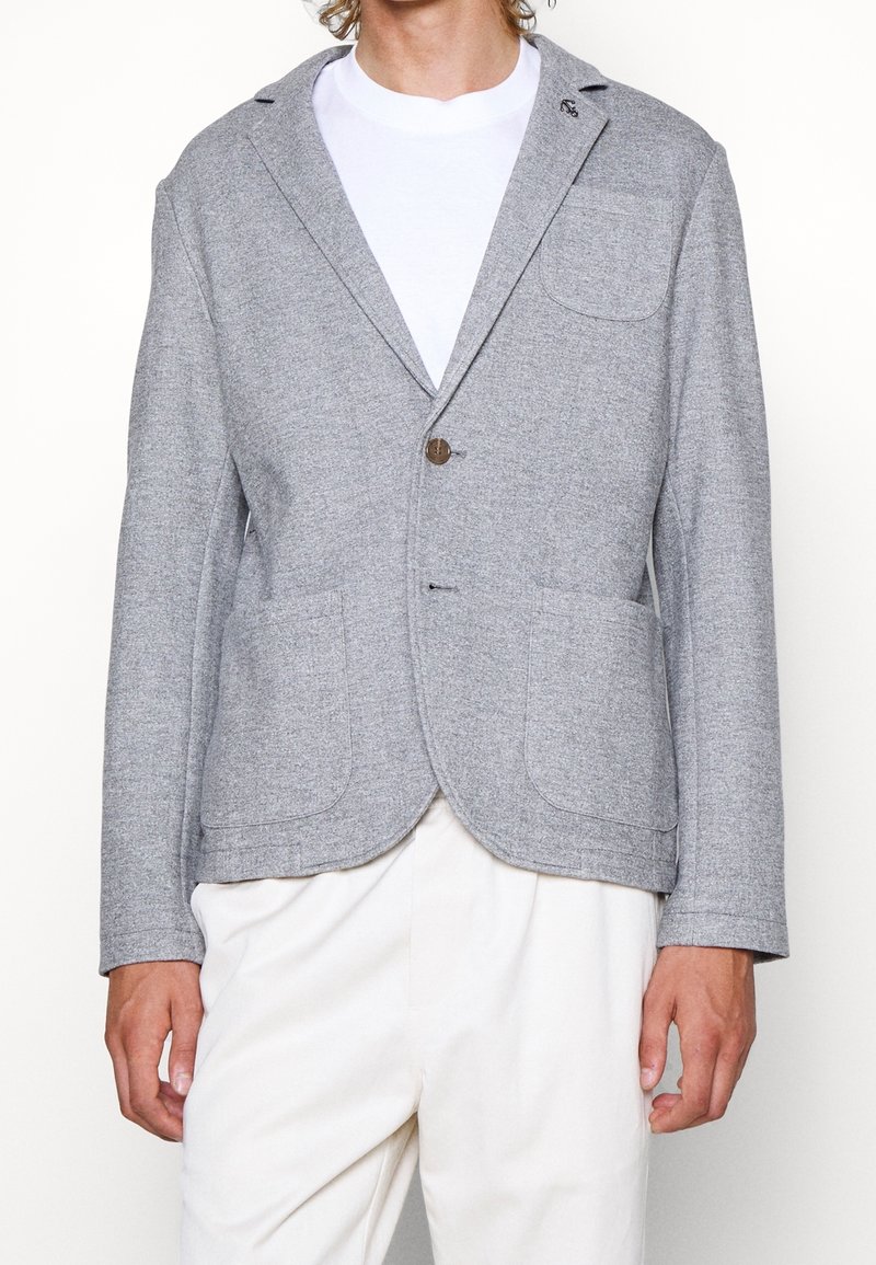 Hombre con blazer gris claro con dos bolsillos frontales y un bolsillo en el pecho sobre una camiseta blanca y pantalones blancos.