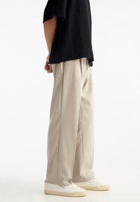 Beige, weite Hosen mit Falten, kombiniert mit weißen Sneakers. Das Oberteil ist ein schwarzes, strukturiertes, locker sitzendes Strickpullover.