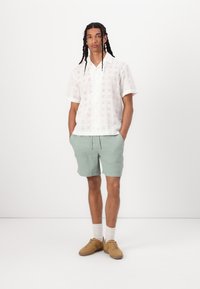 Weißes kurzärmliges Shirt mit Lochstickerei, kombiniert mit hellgrünen gemusterten Shorts und beigen Sneakers, vor einem weißen Hintergrund stehend.