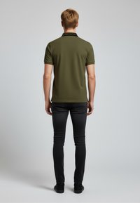 Zelené polo tričko s černým vzorovaným límečkem, doplněné černými úzkými džíny a černými botami. Pohled zezadu ukazuje střih a design outfitu.