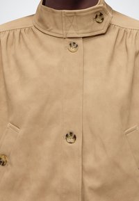 Μπεζ σατέν μπουφάν με ψηλό γιακά, τρία κουμπιά από καβούρι κατά μήκος του μπροστινού μέρους και ραμμένες γωνιακές τσέπες σε κάθε πλευρά.