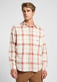 Camisa de cuadros de manga larga en colores crema, rojo y durazno; cuenta con un cuello abotonado, bolsillo en el pecho y una textura de tela suave.