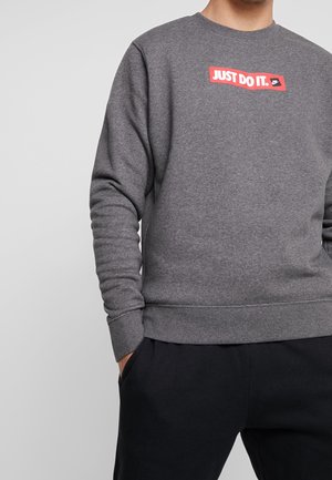 Osoba ubrana w szarą bluzę z czerwoną naszywką "JUST DO IT." oraz czarne spodnie, trzymająca ręce w kieszeniach, na jednolitym, jasnoszarym tle.