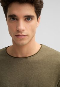 Junger Mann mit lockigem braunem Haar, der ein olivgrünes Rundhals-T-Shirt trägt und direkt in die Kamera schaut, mit neutralem Gesichtsausdruck.