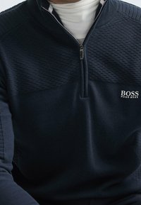 Suéter de media altura azul marino con media cremallera, con textura acanalada, acentos de punto liso y un logotipo blanco en el pecho. Tejido suave con corte clásico.