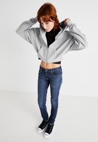 Šedý zipsový cropped mikina cez čierne tričko, skombinovaná s tmavomodrými skinny džínami a čiernymi teniskami s bielymi podrážkami.