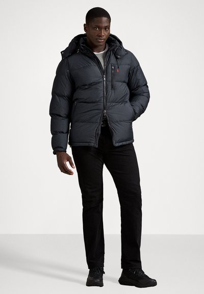 Black puffer jacket with a zippered front and hood, featuring a small logo on the chest. Worn over a light shirt and black pants.
