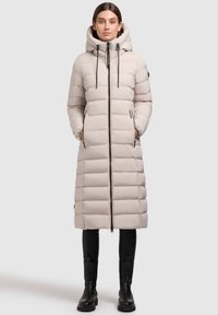 Nafukovacia bunda svetlo béžovej farby s kapucňou, predným zipsom a textúrovaným panelovým dizajnom. Obsahuje dve bočné vrecká a čierne šnúrky na stiahnutie.