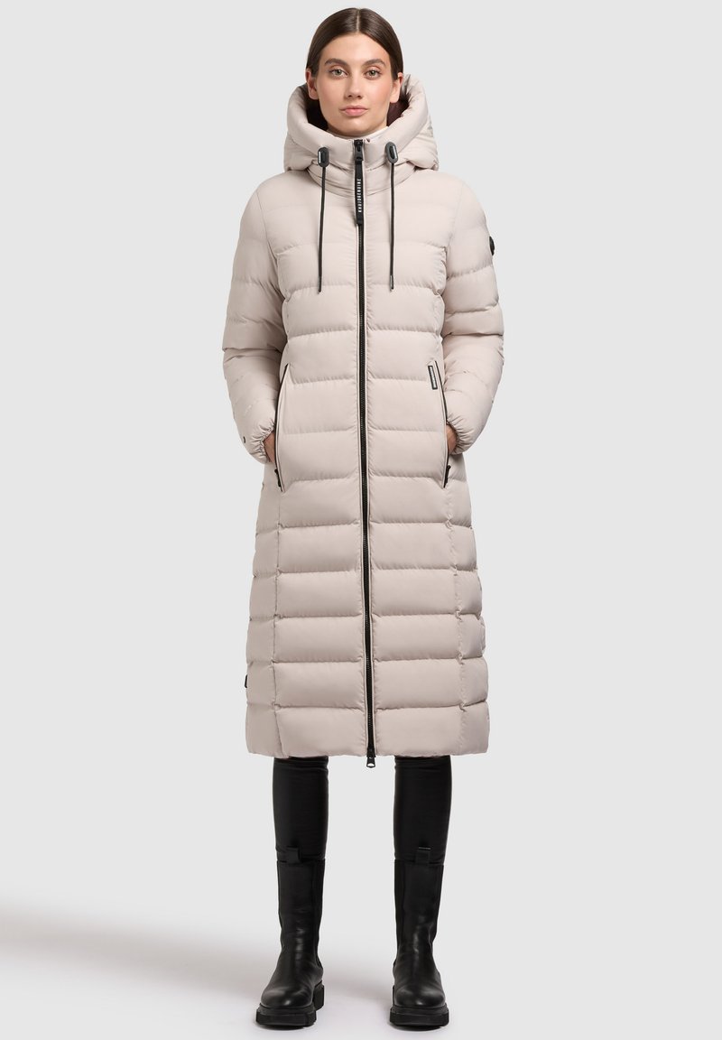 Nafukovacia bunda svetlo béžovej farby s kapucňou, predným zipsom a textúrovaným panelovým dizajnom. Obsahuje dve bočné vrecká a čierne šnúrky na stiahnutie.