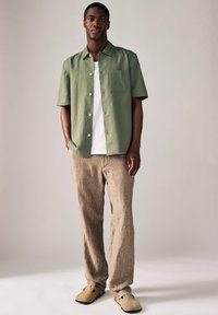 Kortærmet olivengrøn skjorte med en lomme, kombineret med teksturerede beige bukser og lysebrune sko. Neutral baggrund.