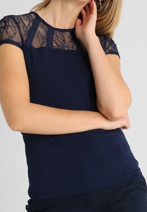 Žena v temně modrém topu s krajkovými krátkými rukávy a horní částí prsou, pózující s jednou rukou dotýkající se krku a druhou rukou zkříženou.