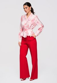 Femme aux cheveux noirs attachés en queue de cheval, portant un chemisier floral rose clair avec une ceinture et un pantalon large rouge vif, posant avec une main dans la poche.