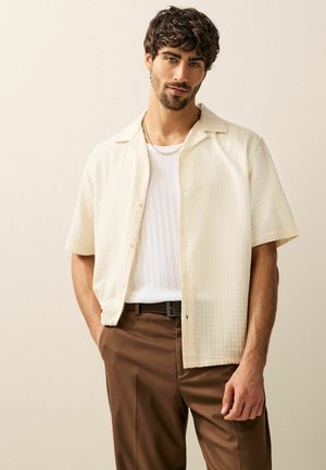 Camisa de botones de manga corta en color crema con textura, llevada sobre una camiseta de tirantes blanca de canalé, combinada con pantalones marrones. Cuello simple y colores neutros.