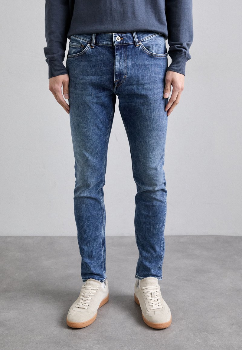 Slim-fit blue denim jeans with a faded finish, five pockets, and button closure, paired with light-colored sneakers on a gray background.
