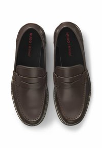 Bruine leren instappers met een gladde textuur, voorzien van een verhoogd naaddesign en contrasterende witte stiksels, met het label "HUGO" in het rood aan de binnenkant.