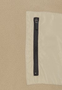 Béžová fleece látka s klopovou kapsou. Kapsa je opatřena černým zipem. Viditelné švy podél okrajů. Celkově hladká textura.