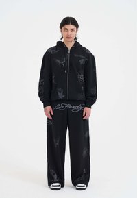 Ung person med mellemlangt hår iført sort zip-up hoodie og brede bukser med tatoveringsstil grafikker samt sneakers, stående op ad hvid baggrund.