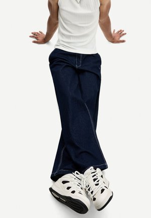 Dark blue wide-leg jeans with white stitching paired with white sneakers featuring black accents and a textured sole.