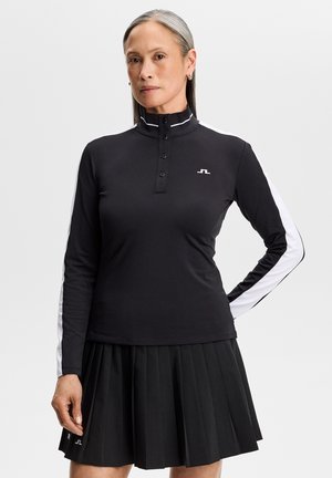 Donna con capelli grigi che indossa un top nero a maniche lunghe con strisce bianche sulle maniche e una gonna nera plissettata, in piedi con una mano sul fianco.