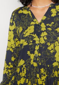 Vestido escuro de tecido com padrão floral amarelo, apresentando uma frente com botões, decote em V e base em camadas. Textura suave, mangas longas.
