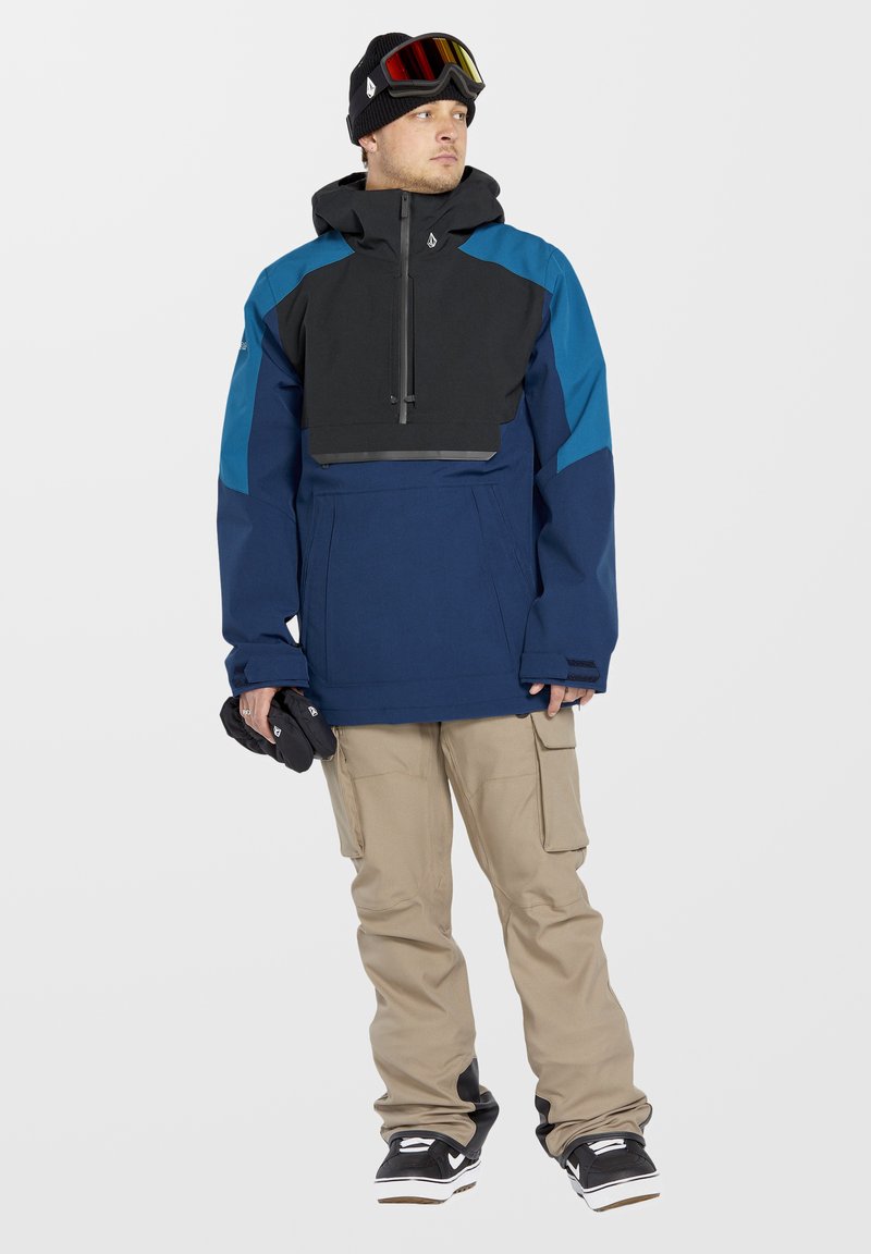 Vandtæt, navy og sort jakke med høj krave, stor frontlomme og justerbar hætte, parret med beige cargo-bukser og sorte sneakers.