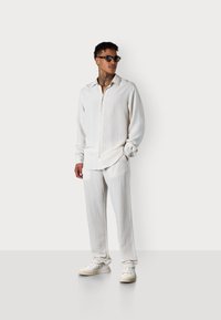 Weißes Leinenhemd und passende Hose mit strukturiertem Finish. Der stehende modell trägt Sonnenbrillen und weiße Sneakers. Minimalistisches Design.