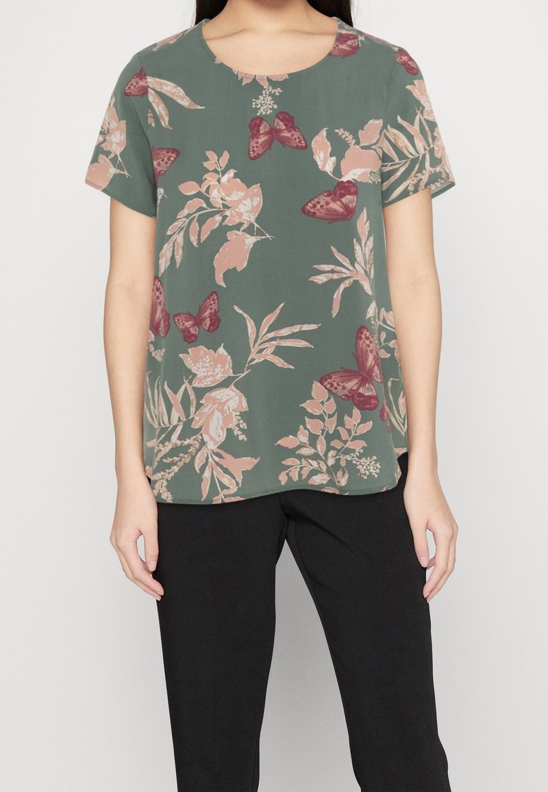 Femme portant un chemisier vert à manches courtes avec des motifs floraux roses et des papillons, associée à un pantalon noir, debout devant un fond uni.