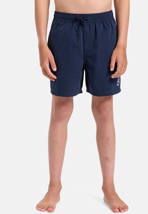 Niño con pantalones cortos de baño azul marino con cintura elástica y logotipo morado en la pierna izquierda, de pie descalzo contra un fondo blanco.