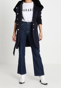 Langer marineblauer Mantel mit silbernen Knöpfen, kombiniert mit einem weißen T-Shirt mit schwarzem Text, hoch taillierten blauen Schlagjeans und glänzenden weißen Ankle Boots.