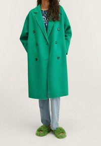 Grønt oversized dobbeltbrystet uldfrakke med sorte knapper, parret med lyseblå jeans og fuzzy grønne slippers, med en afslappet pasform.