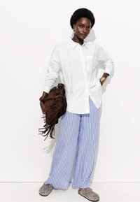 Chemise blanche à boutons, pantalon large rayé violet, sac en daim marron avec des franges, et des chaussures à enfiler en imprimé léopard.