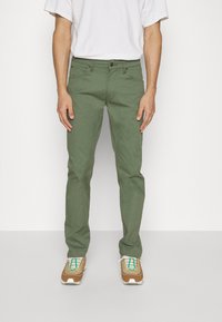 Pantalons en coton verts avec une coupe slim, dotés de cinq poches, d'une couture contrastante et d'une fermeture éclair. Associés à des baskets marron et beige.