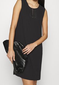 Robe sans manches noire avec un encolure ronde mettant en avant une ligne verticale d'accent en clous. Associée à une pochette noire texturée.