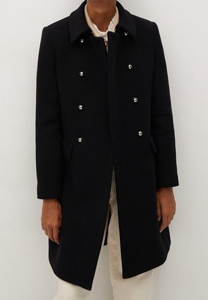 Manteau noir croisé à double boutonnage avec des boutons argentés, porté sur un pantalon beige et un haut blanc, mains détendues sur les côtés, sur un fond uni.