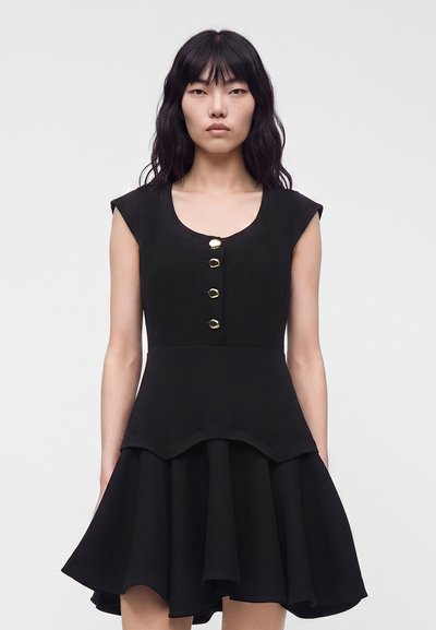 Femme aux longs cheveux noirs portant une robe noire sans manches avec des boutons dorés et une jupe évasée, debout devant un fond clair uni.